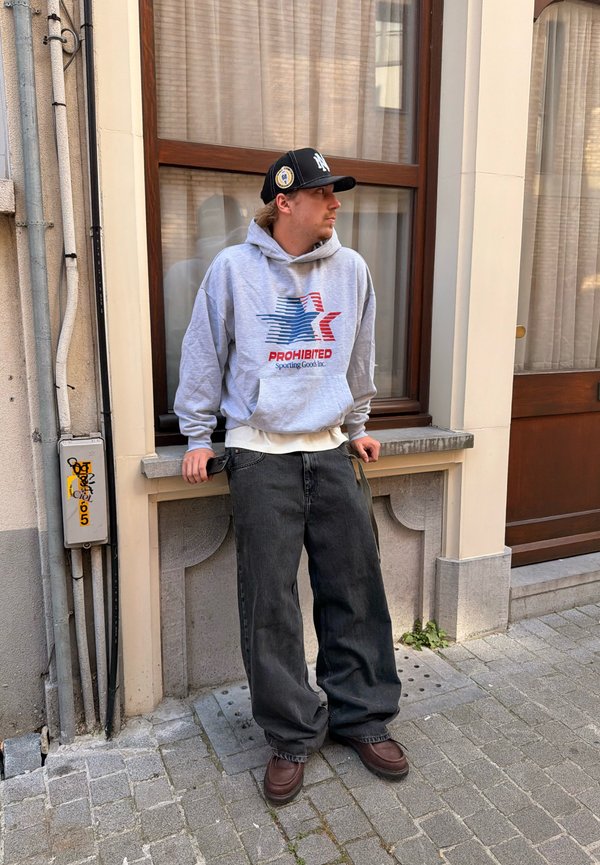 Jeune homme en sweat à capuche gris et casquette noire s’appuyant sur un rebord de fenêtre en pierre dans une rue pavée d’un quartier urbain.