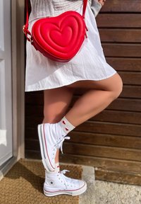 Röd hjärtformad crossbody-väska, vit kjol, vita hög-top sneakers och krämfärgade strumpor med röda hjärtan. Texturerade ytor och kontrasterande färger.