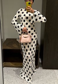 Abito bianco a maniche lunghe con grandi pois neri. Silhouette aderente che si allarga all'orlo. Tiene una piccola borsa rosa chiaro con dettagli dorati.