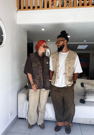 Two people stand indoors wearing animal print clothing and beige outerwear, both with glasses, in a modern living room setting.