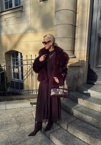 Burgundy fuskappa över en lång, flödande brun klänning. Bär mörka, högklackade stövlar och håller en liten brun handväska. Solbelysta stentrappor.