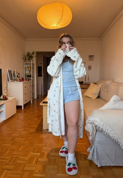 Mujer joven en pijama, con pantuflas de cereza y bata estampada, bebe de una taza en una sala de estar acogedora con suelo de madera y luz suave.