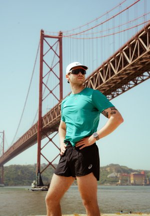 Uomo atletico in una maglietta teal traspirante e pantaloni corti neri posa all'aperto, con un ponte sospeso rosso sullo sfondo.