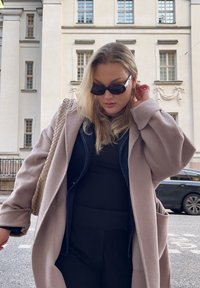 Šviesiai rožinė per didelė striukė, juodas viršus ir kelnės, su tekstūruotu pintu dideliu krepšiu. Tams�ūs akiniai saulėms pabrėžia aprangą.