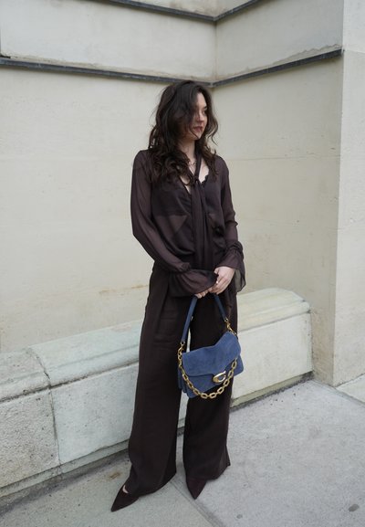 Mujer con cabello largo y oscuro que lleva una blusa negra translúcida y pantalones de piernas anchas, sosteniendo un bolso azul con cadena dorada, de pie frente a una pared pálida.