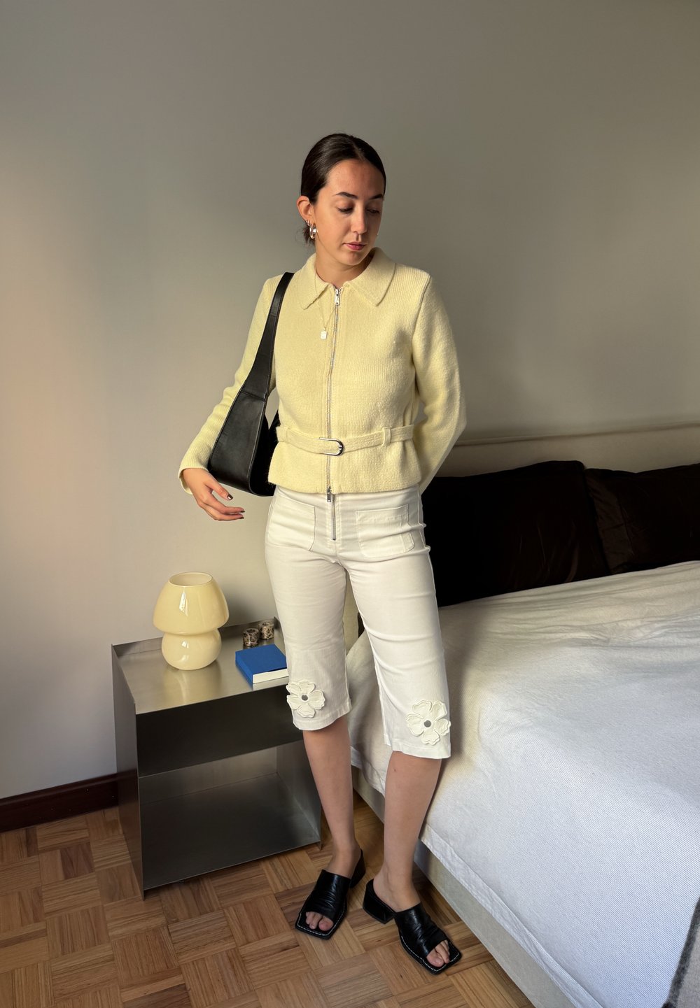 Jachetă galbenă cu fermoar și guler, pantaloni albi scurți cu broderie florală, sandale plate negre și o geantă de umăr neagră. Fundal în tonuri neutre.