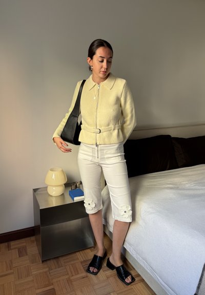 Chaqueta amarilla con cremallera y cuello, pantalones blancos cortos con bordado floral, sandalias planas negras y un bolso negro de hombro. Fondo en tonos neutros.