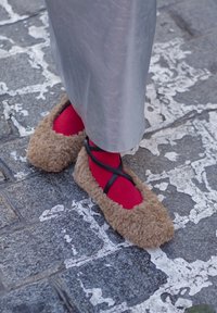 Φουντωτές μπεζ μπαλαρίνες με μαύρες διασταυρούμενες λωρίδες, φορεμένες πάνω από κόκκινες κάλτσες, να στέκονται σε μια υφασμάτινη γκρι πλακόστρωτη επιφάνεια.