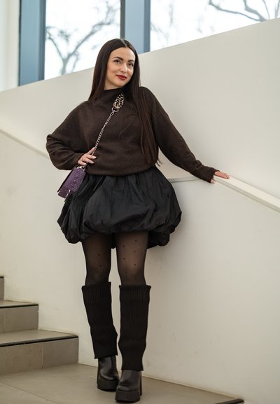 Mujer con cabello largo y oscuro, vestida con un suéter marrón, falda negra, medias con estampado de corazones, botas y un bolso cruzado morado, de pie junto a unas escaleras.