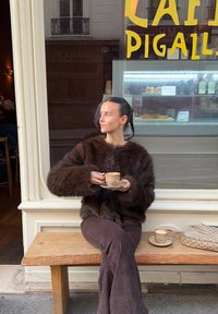 Suéter de piel sintética marrón, pantalones de pana marrón, sosteniendo una taza de café en un banco de madera frente a un café con carteles amarillos.