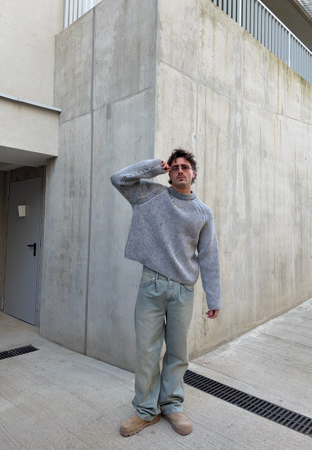 Grå randig tröja, ljusblå baggy jeans, bruna stövlar, står mot en texturerad betongvägg. Minimalistiska accessoarer och kort hår.
