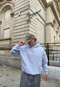 Grå oversize hættetrøje med frontlomme, kombineret med mørkegrå, løstsiddende bukser. En olivengrøn kasket med hvidt logo. Urbant miljø.