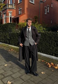 Um homem está a pé numa passadeira pavimentada, vestindo um sobretudo preto, um cardigã cinzento, uma camisa branca, uma gravata preta e calças largas pretas realçadas por botas pretas.