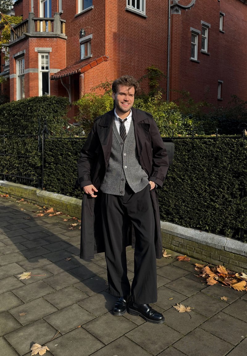 Un uomo si trova su un sentiero lastricato, indossando un trench nero, un cardigan grigio, una camicia bianca, una cravatta nera e ampi pantaloni neri abbinati a stivali neri.