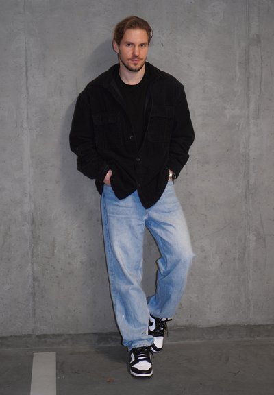 Chaqueta de pana negra sobre una camisa negra, jeans holgados color azul claro y zapatillas negras y blancas. El fondo es una pared de textura gris.