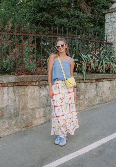 Top sin mangas azul, falda larga blanca con patrones coloridos, bolso cruzado amarillo claro y zapatillas azules. Entorno al aire libre con vegetación.