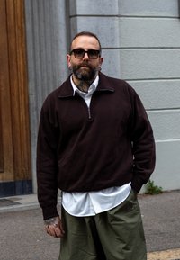 Sweatshirt brun zippé sur une chemise blanche à col, associé à un pantalon vert à jambes larges. La personne a des tatouages et porte des lunettes de soleil noires.