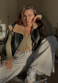 Schwarze Lederweste mit Schaffellkragen, gerippter olivgrüner Cropped-Top, hellbeige weit geschnittene Hose und graue Sportschuhe mit Akzenten.