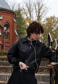 Sort læderjakke med en struktureret finish, der har subtil branding. Mørke bukser og tilbehør inkluderer et bælte og hovedtelefoner. Betontrapper og efterårsblade i baggrunden.