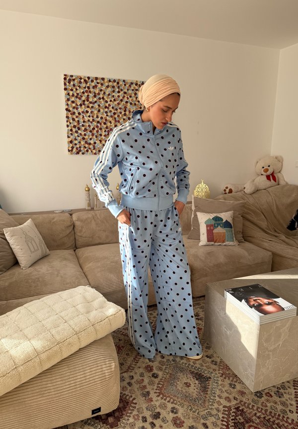 Femme portant un survêtement bleu clair à pois et un foulard beige sur la tête, debout dans un salon avec un canapé beige, des coussins, un ours en peluche et un tapis à motifs.
