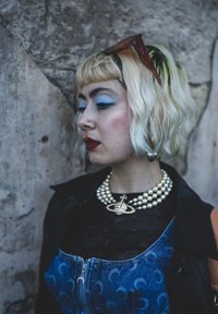 Jeansklänning med dragkedja, lager-på-lager över en genomskinlig svart topp. Pärlhalsband med en stjärna och en planet som hängsmycke. Kort, vågigt hår med gröna toppar.