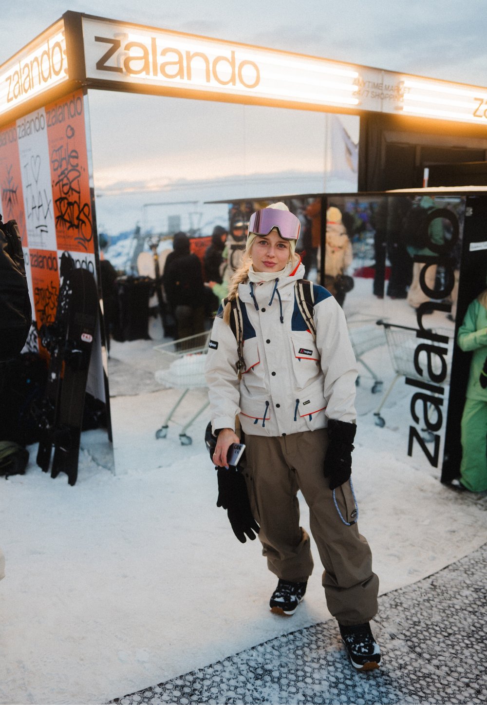 Jeune femme en tenue de ski et lunettes de protection se tient sur la neige près d'un stand éclairé Zalando, avec des personnes et des caddies en arrière-plan.