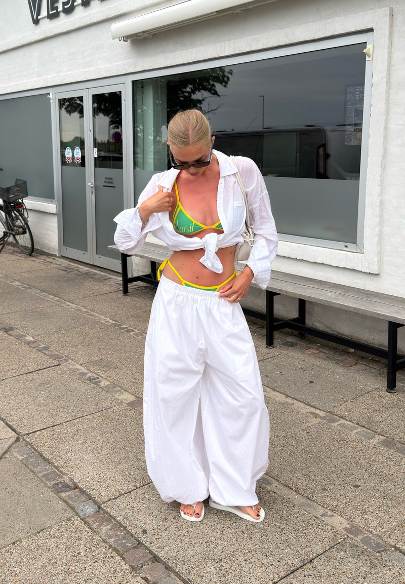 Ženska, oblečena v rumeno-zeleni bikini pod vezano belo srajco in široke bele hlače, stoji na pločniku blizu stavbe z klopco.