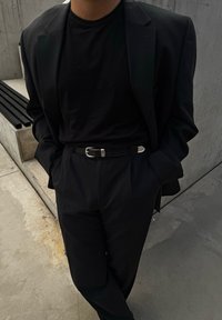 Μαύρο σακάκι κομμένο στα μέτρα πάνω από ένα μαύρο t-shirt, με μια λεπτή μαύρη ζώνη με ασημένια αγκράφα. Στηρίζεται σε έναν τσιμεντένιο τοίχο.