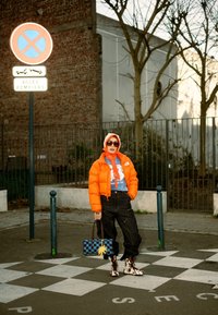 Personne portant une veste matelassée orange, un sweat à capuche, un pantalon noir, des bottes à motifs et des lunettes de soleil, tenant un sac à main bleu à carreaux sur un passage pour piétons à carreaux.