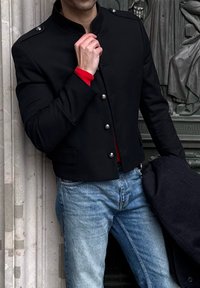 Jachetă neagră în stil militar, cu guler înalt, nasturi argintii și croială mulată. O cămașă roșie este vizibilă dedesubt. Blugii albaștri completează ținuta.