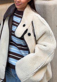 Femme portant un manteau sherpa crème avec des boutons et bordures noirs, par-dessus un pull rayé bleu, noir et blanc, debout contre un mur en pierre.