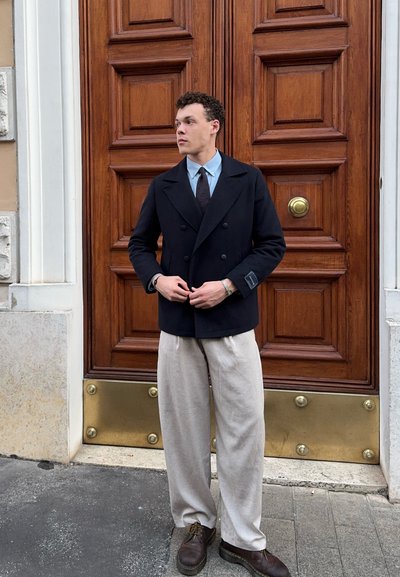Blazer azul marino de doble botonadura, camisa de color azul claro, corbata marrón, pantalones beiges y zapatos marrones; de pie junto a una puerta de madera con acentos de latón.