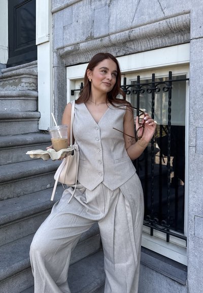 Chaleco de lino gris claro y pantalones de pierna ancha con pliegues. Sosteniendo una taza de café y un estuche para gafas de sol, de pie en escalones de piedra.