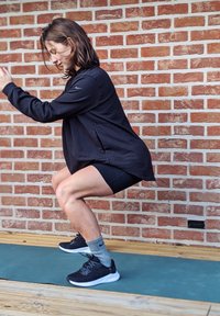 Un sweat-shirt de sport noir et un short portés par une personne effectuant un squat sur un tapis vert devant un mur en briques ; des chaussures de course noires et des chaussettes grises.