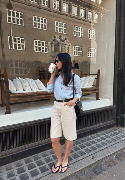 Camisa de botones azul claro, pantalones cortos beige, cinturón negro y chanclas. Bolsa negra con herrajes dorados. Edificio reflejado en el fondo.