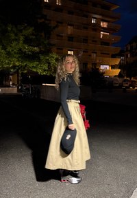 En person har på seg en svart langermet skjorte, et flytende beiges skjørt og sølvfargede joggesko, og holder en svart veske med et dekorativt accent.