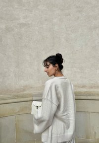 Un pull surdimensionné gris clair avec des manches larges, un tricot texturé et un ourlet légèrement côtelé. Tenant un petit sac à main blanc. Fond simple.