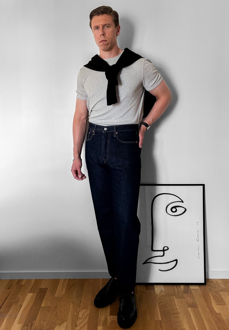 Homme en t-shirt gris, pull noir sur les épaules, jeans foncés et chaussures noires, debout à côté d'une œuvre d'art abstraite minimaliste encadrée représentant un visage sur un sol en bois.