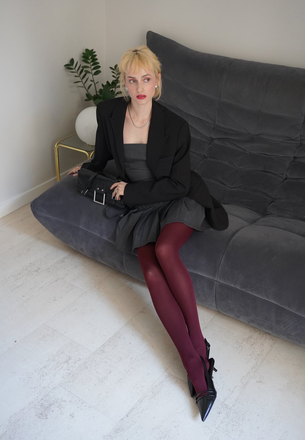 Blazer noir sur une robe grise, collants rouges et escarpins noirs. Le canapé est gris avec une surface texturée. Un petit sac à main noir est présent.