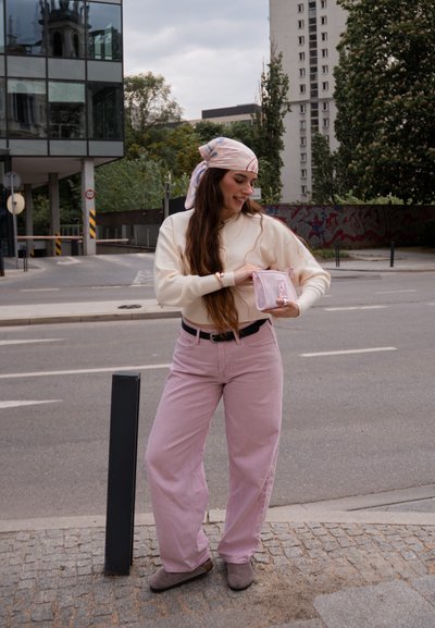 Joven mujer con pañuelo rosa en la cabeza, suéter crema y pantalones rosas, sosteniendo una pequeña bolsa rosa de malla en una acera de la ciudad cerca de la calle.