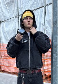 Fleece de ganga preto com fecho éclair e mangas oversized, utilizado sobre uma camisa branca. Segura um haltere, com um gorro amarelo com detalhes azuis.