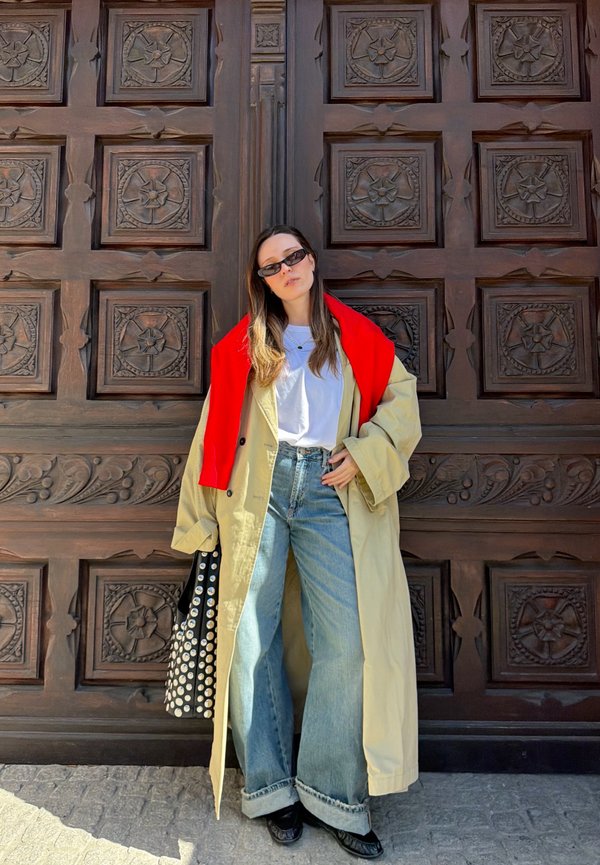 Kvinde iført beige trenchcoat med rød sweater draperet over skuldrene, hvid skjorte, brede jeans og solbriller, stående ved udsmykkede udskårne trædøre.