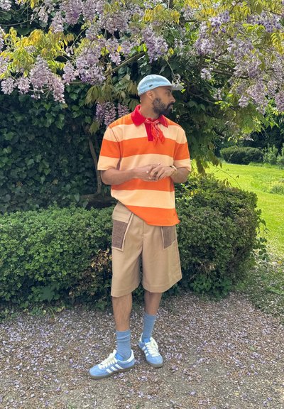 Polo de rayas de manga corta en colores naranja y crema, con cuello rojo, pantalones cortos caqui con detalles en los bolsillos, zapatillas azules con cordones blancos y calcetines azules.