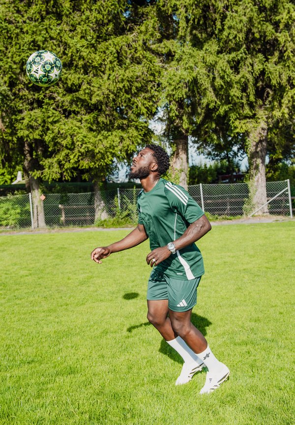 Mann in grüner Sportbekleidung bereitet sich darauf vor, einen Fußball auf einem grasbewachsenen Feld mit hohen Bäumen und einem Zaun im Hintergrund zu köpfen.