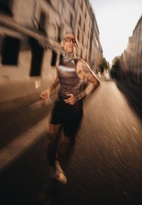 Sivi atletski majica bez rukava, crne kratke hlače i bijele čarape. Ima tetovaže na rukama, pozicija trčanja s mutnom pozadinom, što dočarava pokret.