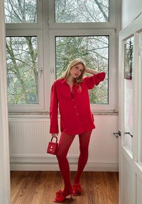 Chemise rouge à boutons, collants assortis et sandales avec des nœuds. Petit sac à main rouge. Intérieur avec de grandes fenêtres et un parquet en bois.