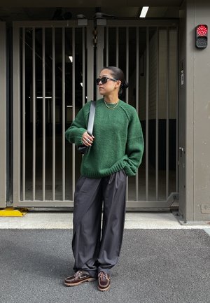 Grüner gestrickter Pullover, lockere schwarze Hose und braune Bootsschuhe. Steht vor einem Parkhaus mit vertikalen Metallstangen.