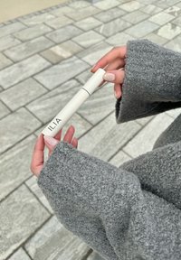 Προϊόν ILIA σε κομψό, κυλινδρικό λευκό σωλήνα με μια μαύρη γραμμή, κρατιέται από ένα χέρι που φοράει υφασμάτινη γκρίζα ζακέτα σε φόντο πέτρας.