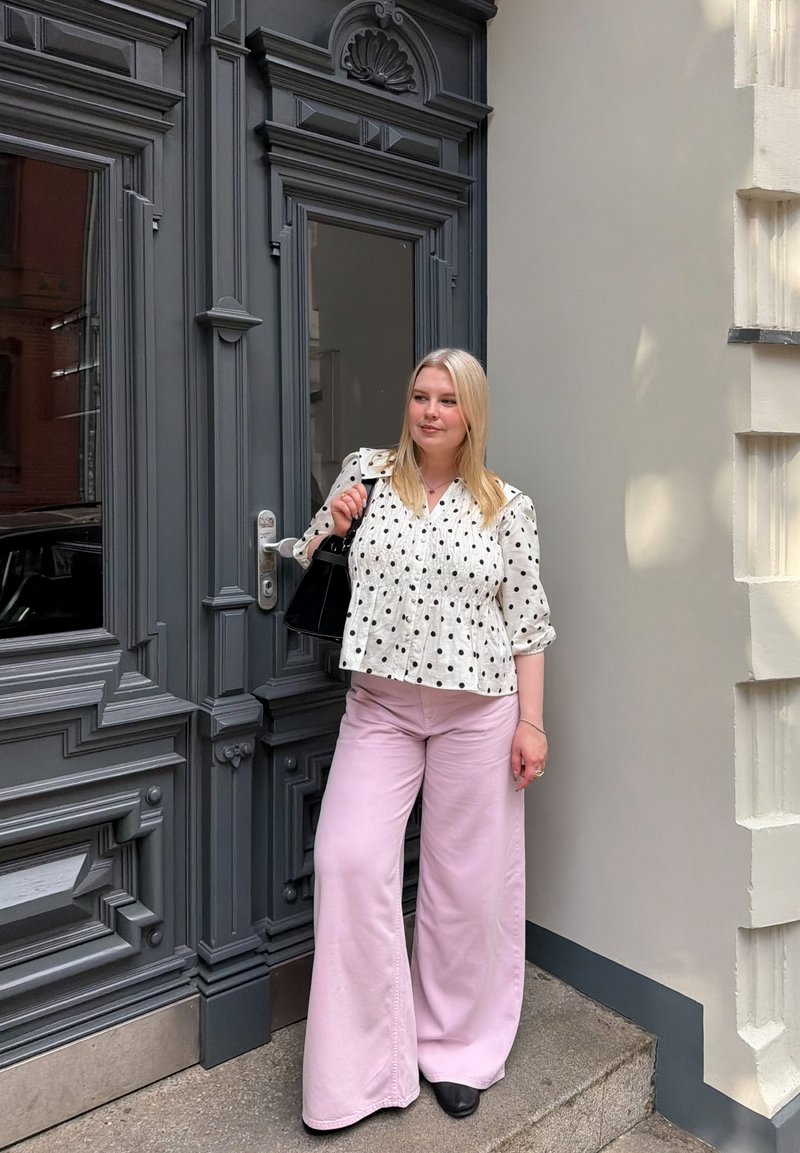 Femme aux cheveux blonds portant un chemisier blanc à pois et un pantalon large rose se tenant près d'une porte ornée gris foncé tenant un sac à main noir.