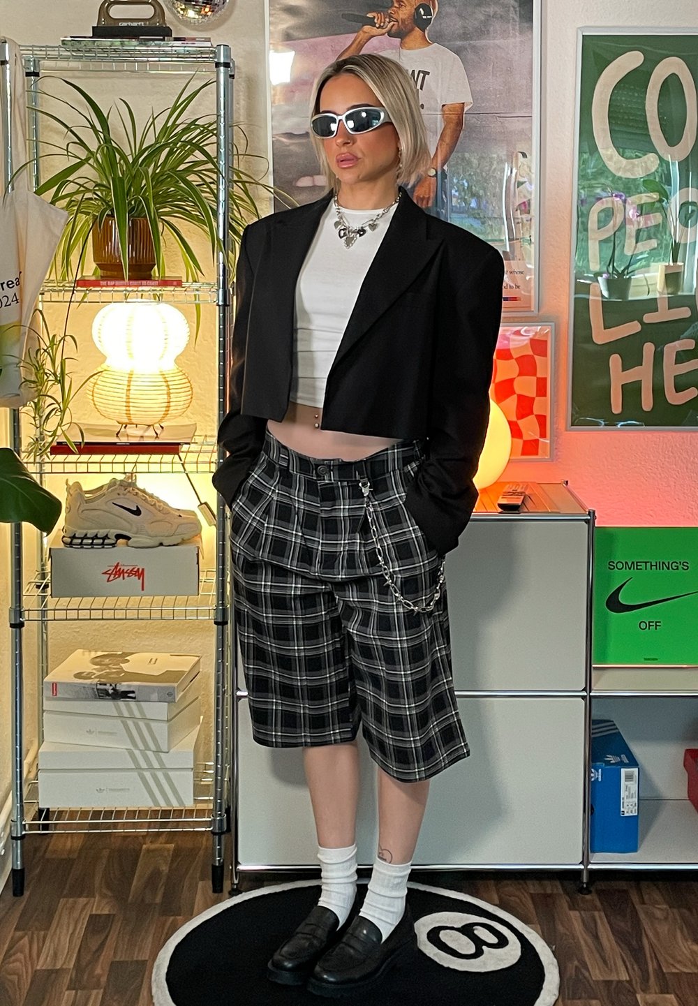 Črn skrajšan blazer preko belega majice, v kombinaciji s črno-belimi kratkimi hlačami s karirastim vzorcem, ki vključujejo verige. Črne čevlje in bele nogavice dopolnjujejo videz.